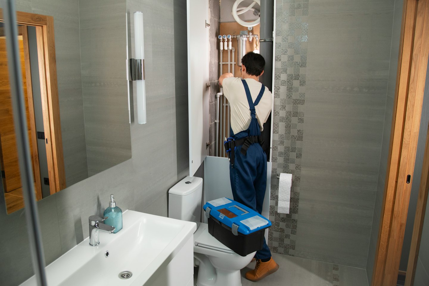 Technician repairs plumbing behind toilet in grey tiled bathroom.